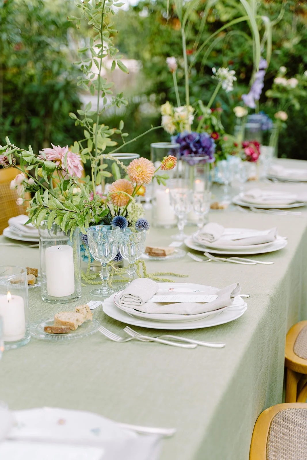 Elegant outdoor dining table decorated with floral arrangements in vases, candles, and place settings with white plates and napkins, set in a garden.