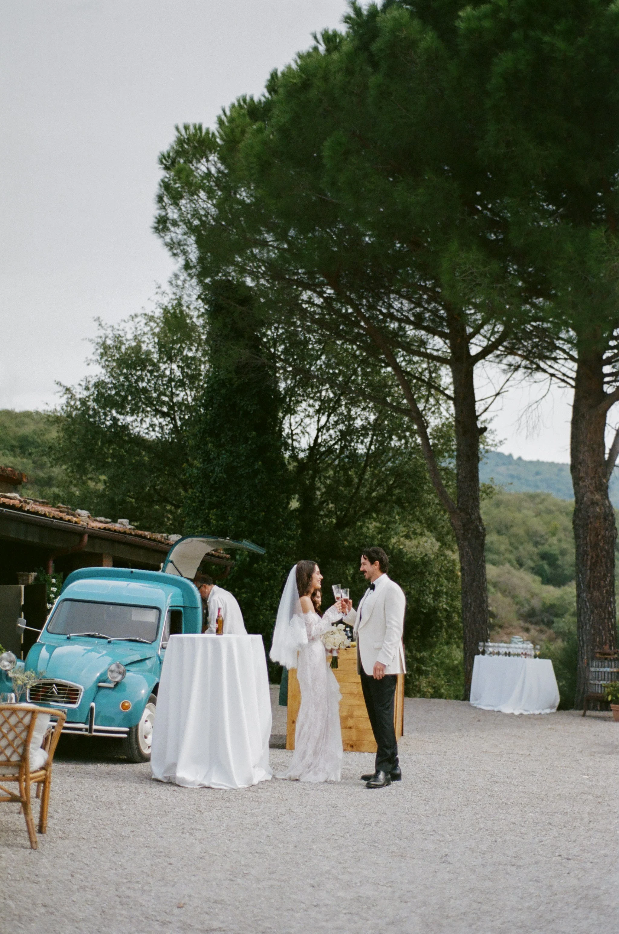 Bride and groom toasting with glasses at outdoor wedding reception with vintage blue car in background.