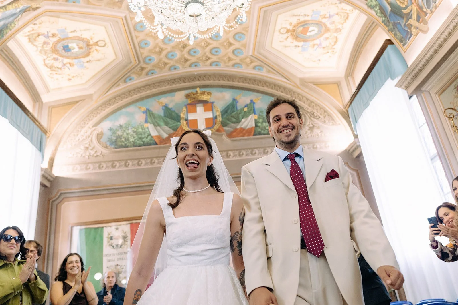 A bride and groom smiling happily during their wedding ceremony in a decorated ornate hall, with guests clapping and taking photos.