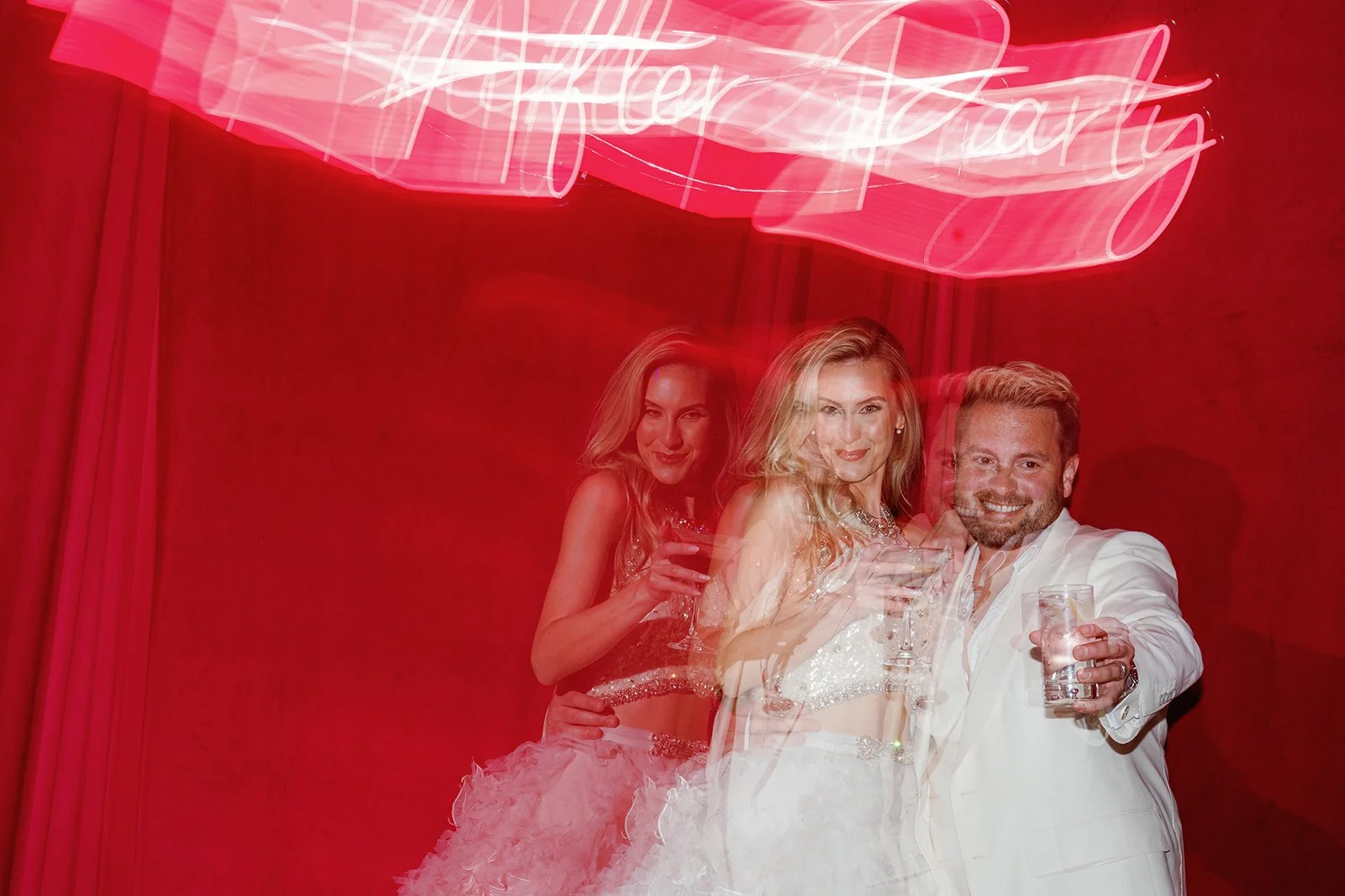 Three people at a celebration holding drinks, with a pink neon sign above them that reads 'Bachelor Party' and red curtains in the background.