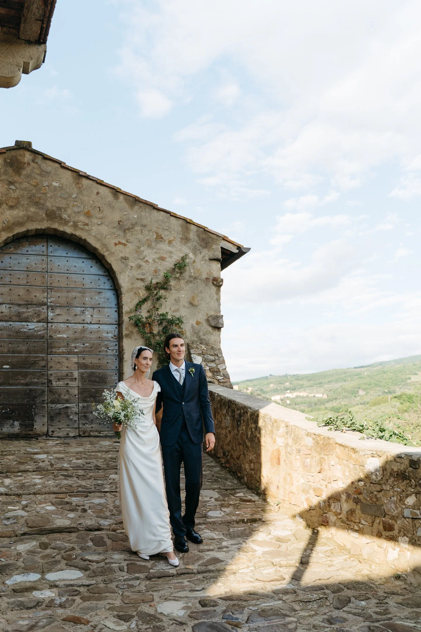A bride and groom walk arm in arm along a stone pathway outside a rustic stone building, with a scenic landscape and sky in the background.