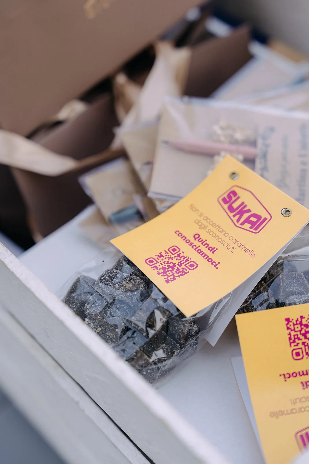 Close-up of a yellow and pink Sukai brand label with QR codes, placed on small dark packaged items inside a plastic bag, with some paper and cardboard packages in the background.