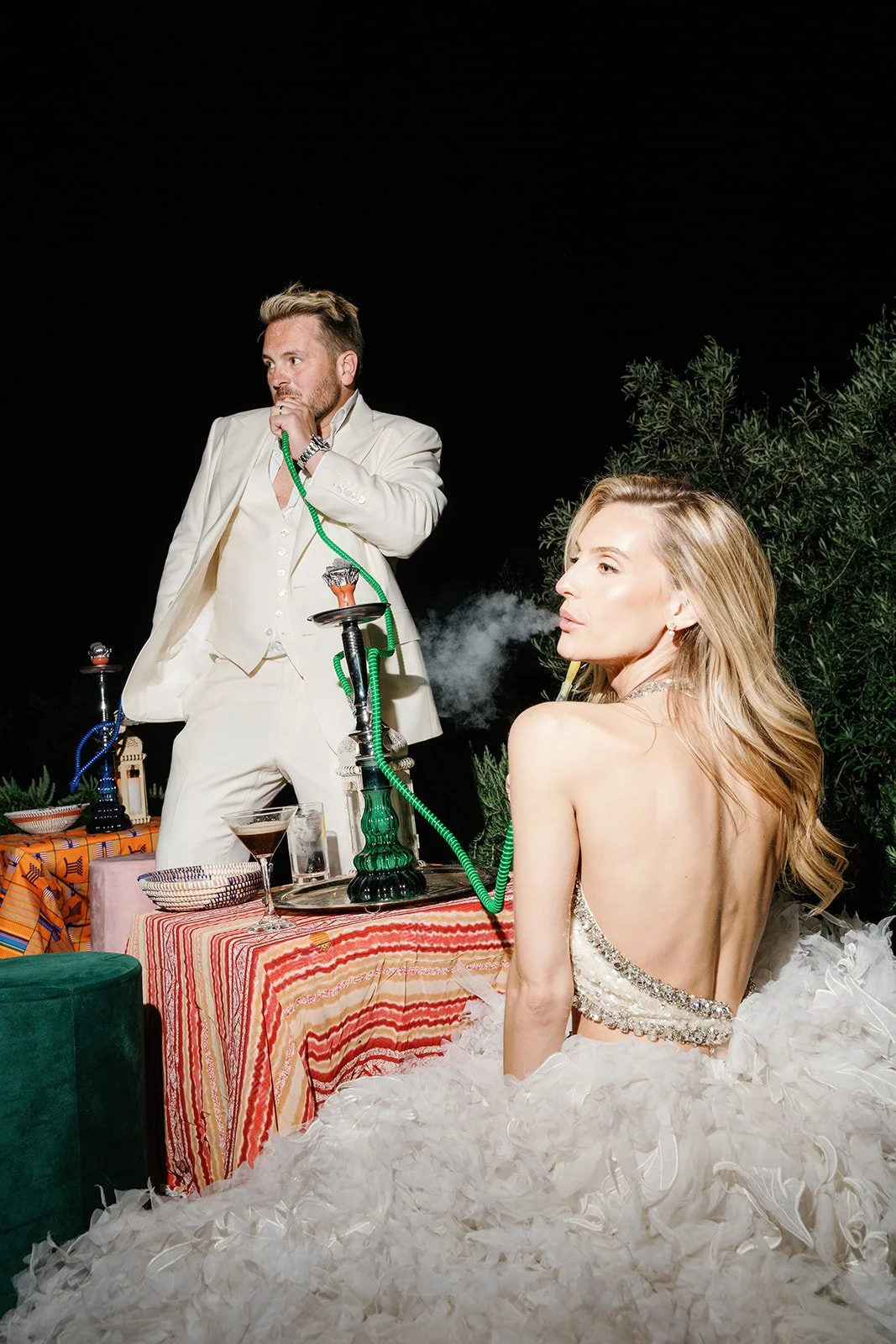A man in a white suit is using a hookah. A woman in a glamorous dress with a low back and feathered skirt is sitting nearby, smoking from a straw connected to the hookah. They are outdoors at night with trees in the background and a multicolored tabl