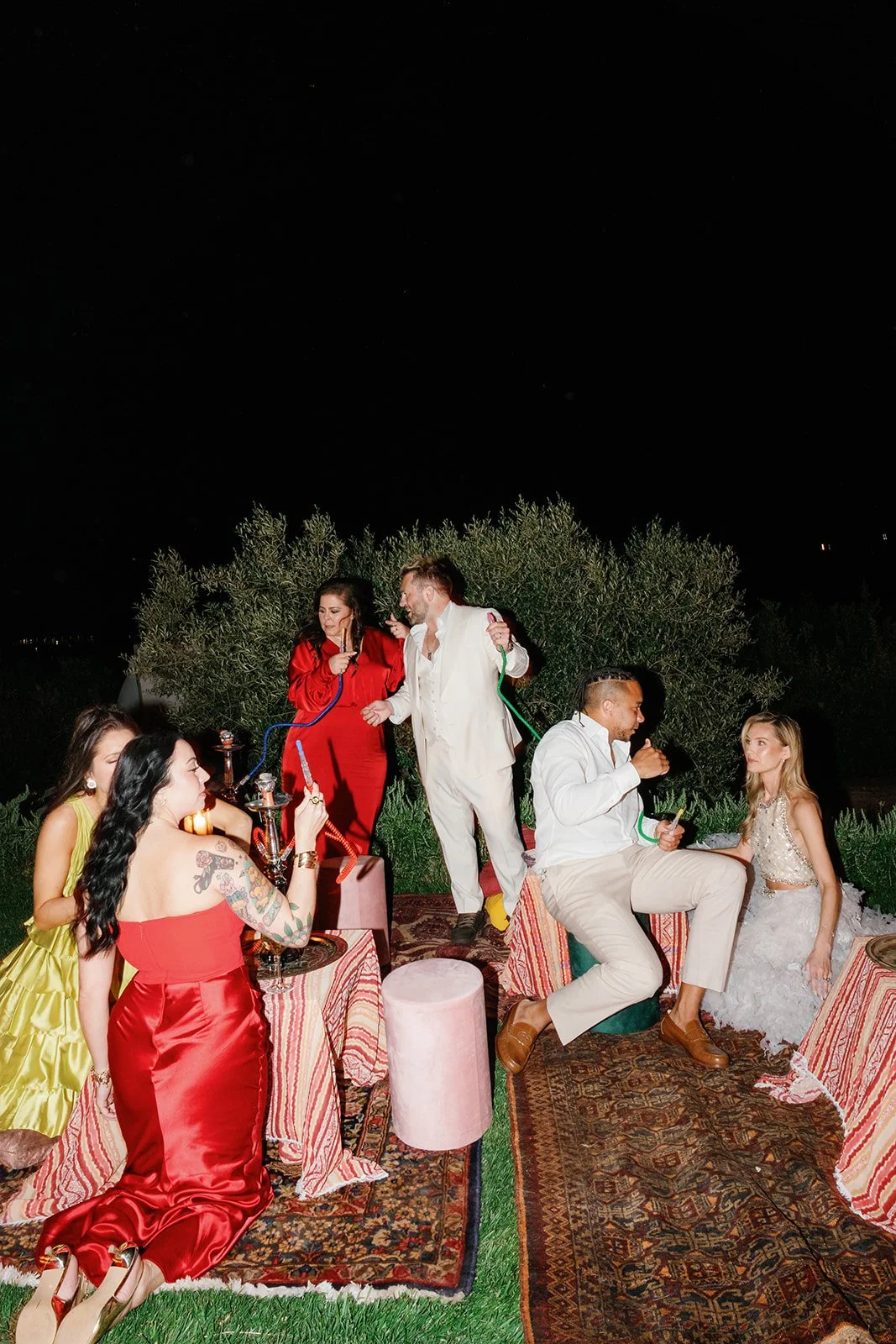 Group of people at a nighttime outdoor gathering, sitting on Persian rugs, using hookahs, and enjoying drinks. Some are standing, and the woman on the right is dressed in a white dress, possibly a wedding gown.