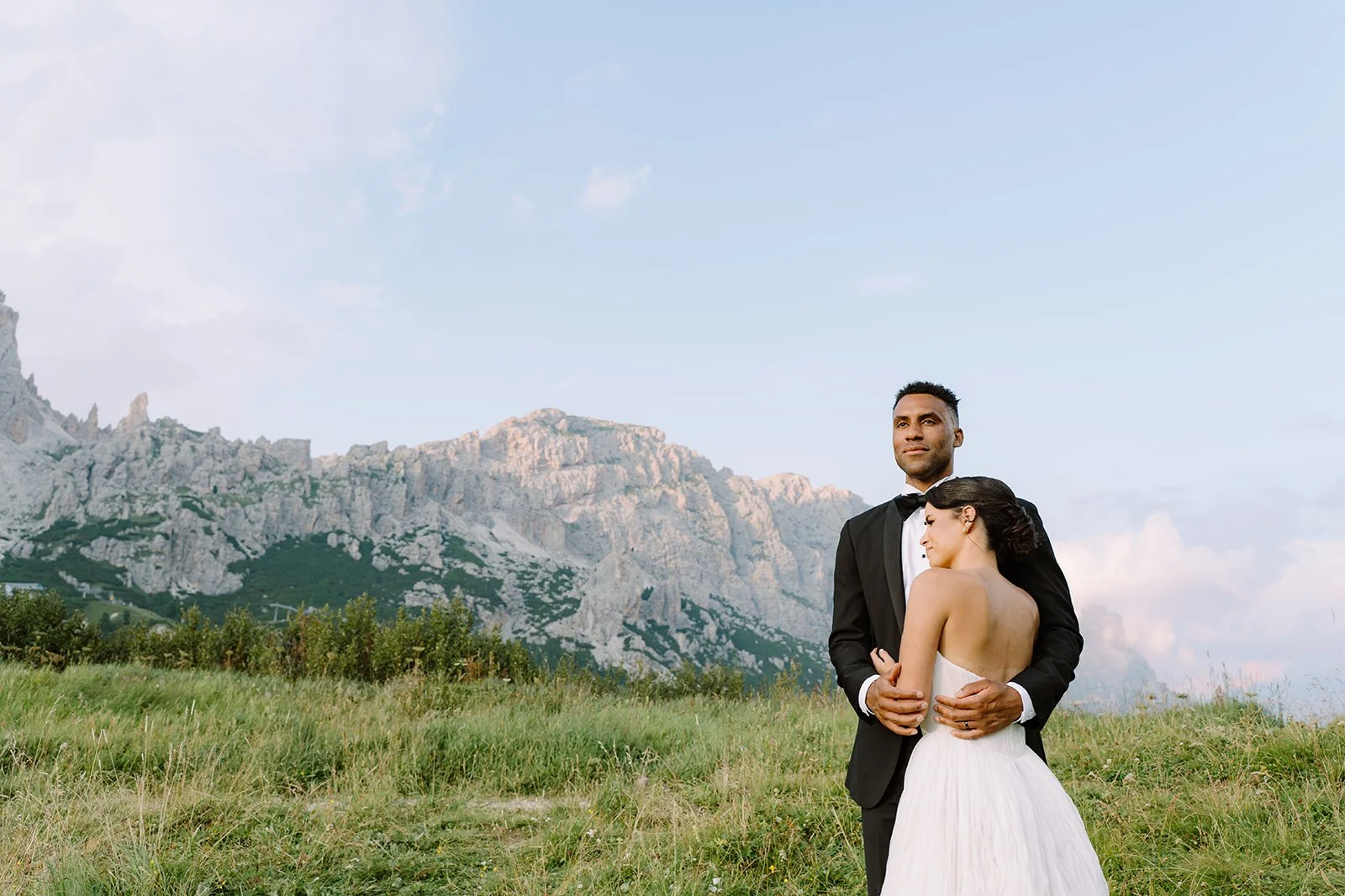 A wedding couple standing in a grassy field with mountains in the background. The groom is in a black tuxedo and the bride in a strapless white dress.