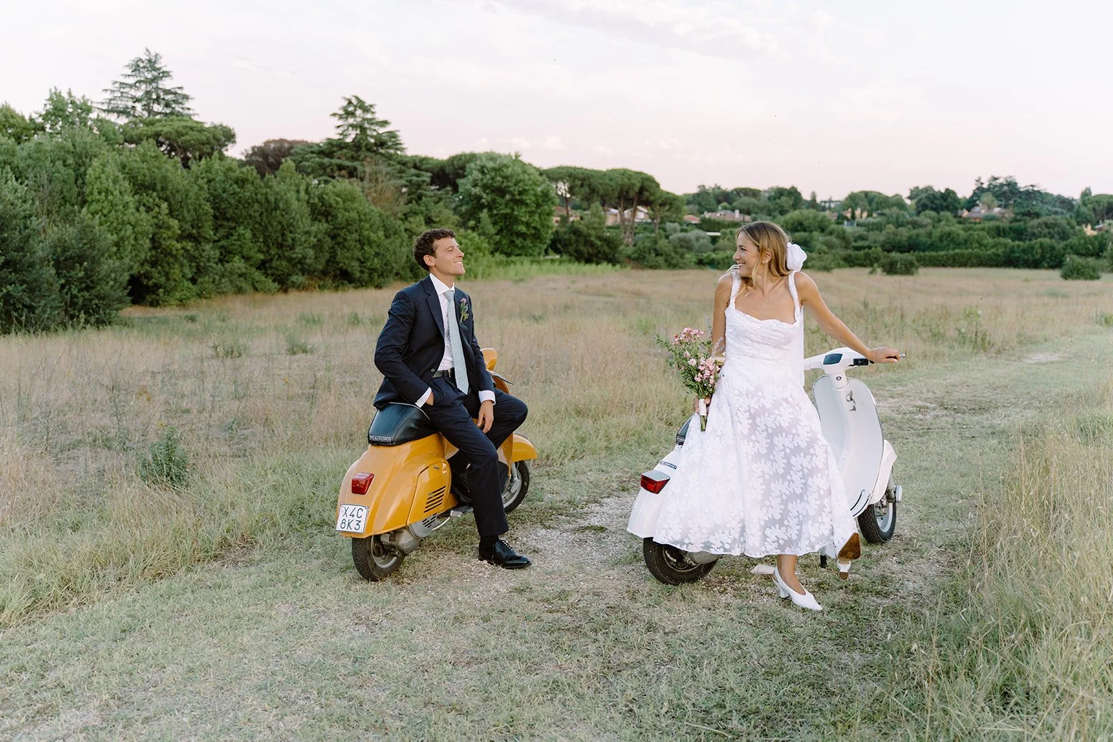 A man in a suit sitting on a yellow scooter and a woman in a white wedding dress standing beside a white scooter in a grassy field with trees in the background.