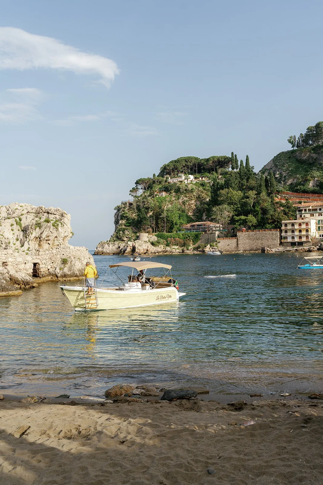 A boat sailing near a rocky shoreline with a people on board, surrounded by a coastal landscape with lush green hills and buildings in the background.