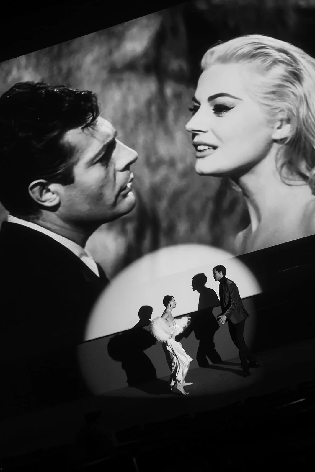 A black and white image of a man and woman looking at each other closely on a large screen, and their shadows are projected onto a wall below.