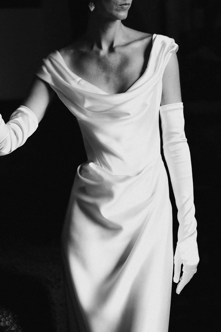 A woman wearing a white dress and long gloves, with diamond earrings, posing in a black and white photo.
