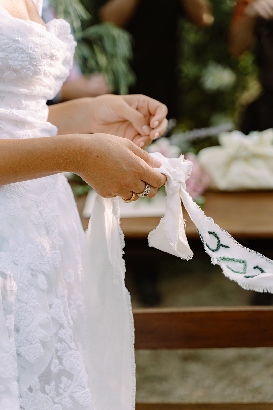 A person wearing a white lace dress holding and tying a cloth ribbon with green embroidery.