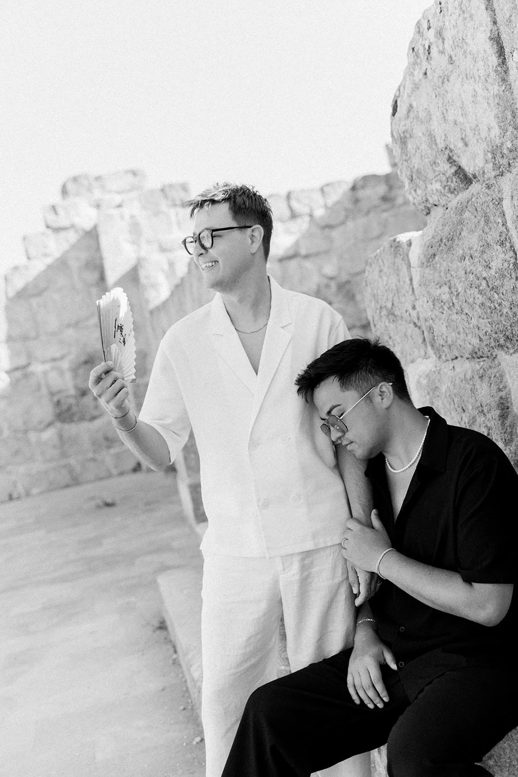 Two young men with glasses and jewelry leaning against a stone wall outdoors, one smiling and holding a fan, the other resting his head on the other's arm and sitting with closed eyes.