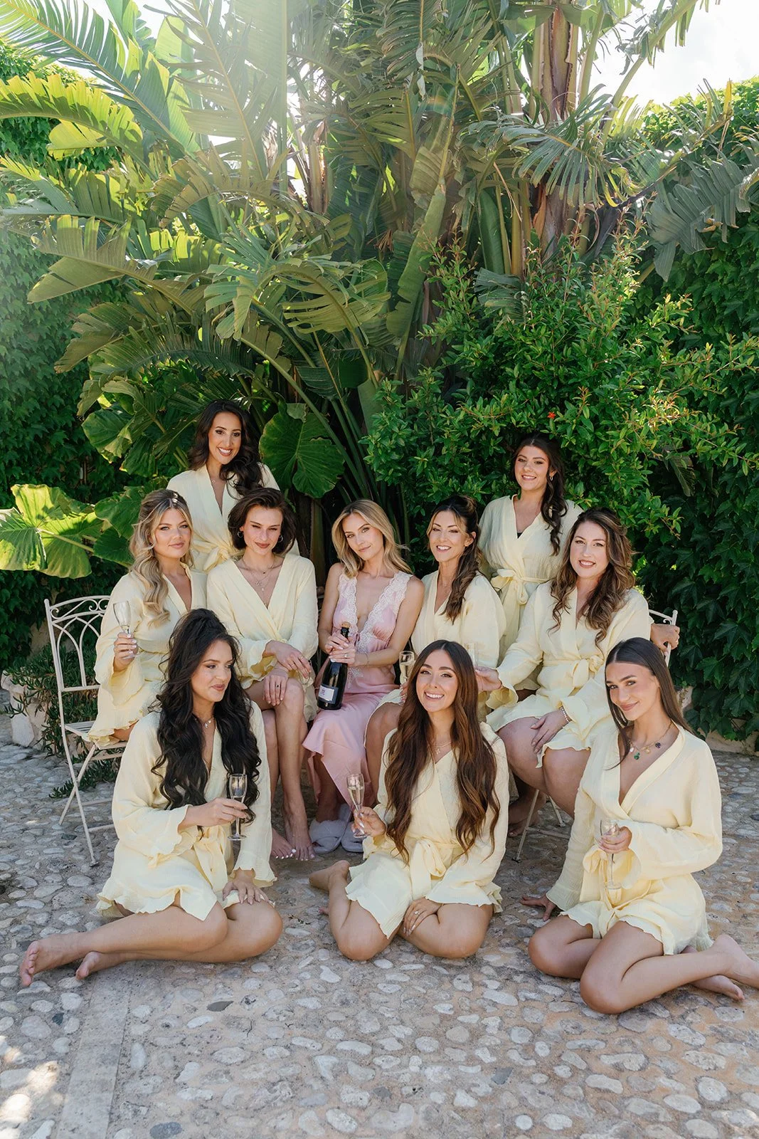 A group of women in pastel-colored robes, some holding glasses of champagne and a bottle, gathered outdoors on a stone patio surrounded by lush green tropical plants, celebrating a special occasion.
