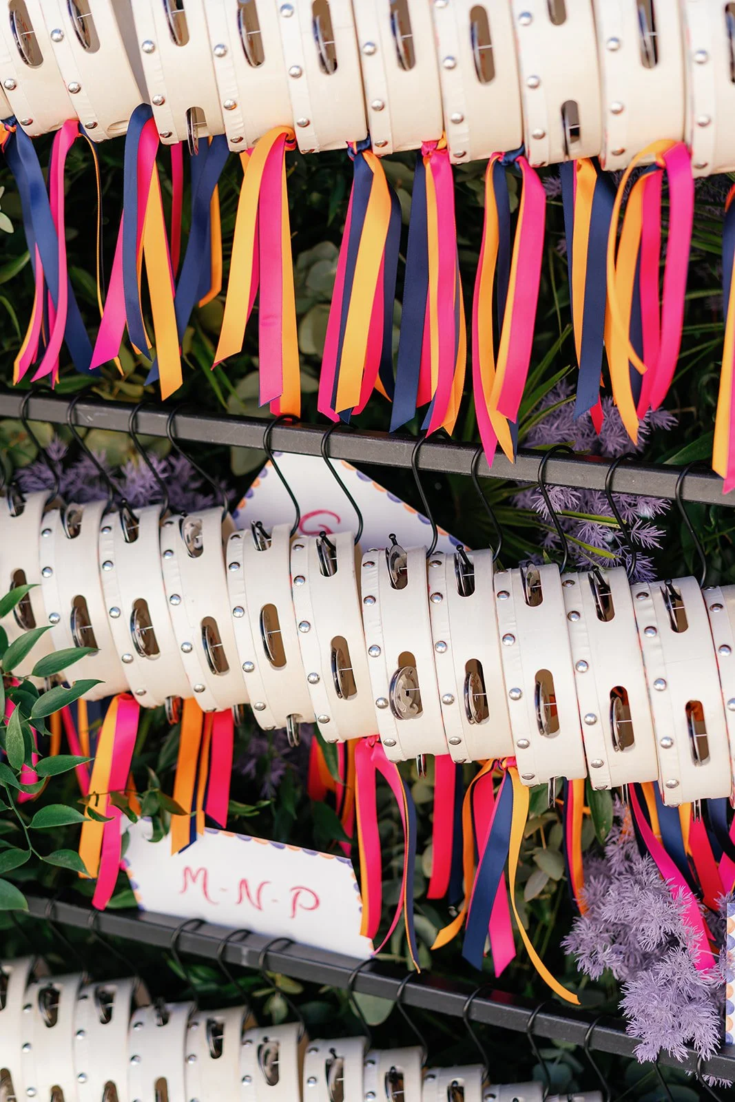 Colorful ribbons hang from white leather or faux leather bags with cut-out handles, displayed on black metal racks, surrounded by purple flowers and green foliage, with a handwritten sign reading 'M-N-P' in red ink.