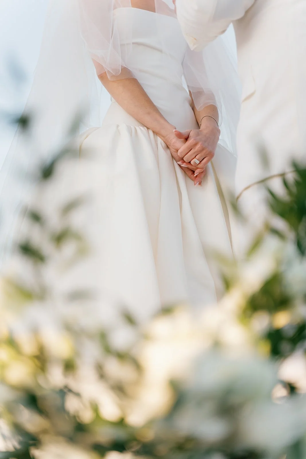 Close-up of a bride in a white wedding dress, standing with her hands clasped in front of her, showing her wedding ring, with blurred white flowers in the foreground.
