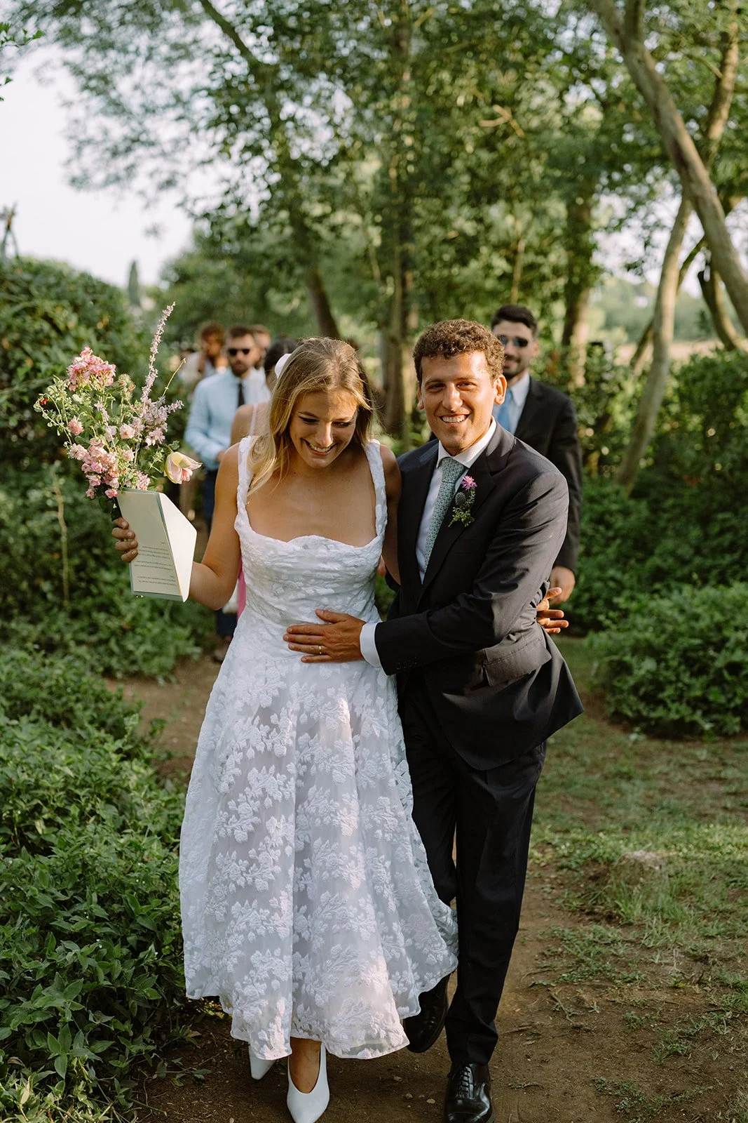 A newlywed couple is walking outdoors on a trail, smiling and enjoying their wedding day; the bride holds a bouquet of pink flowers, and the groom is holding her around the waist.