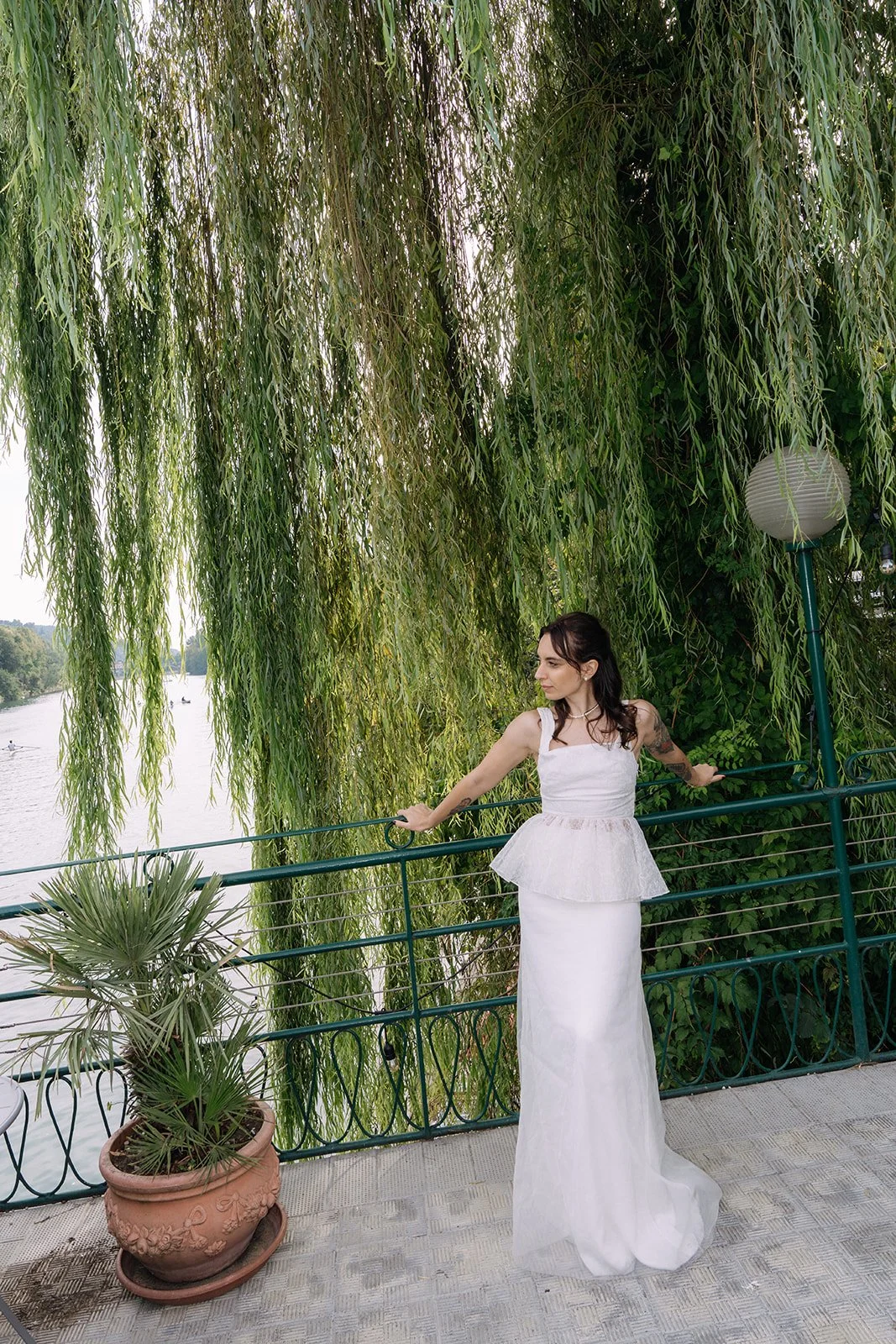 Woman in a white dress standing on a riverside balcony with hanging willow trees and potted plant around, holding onto a green railing.