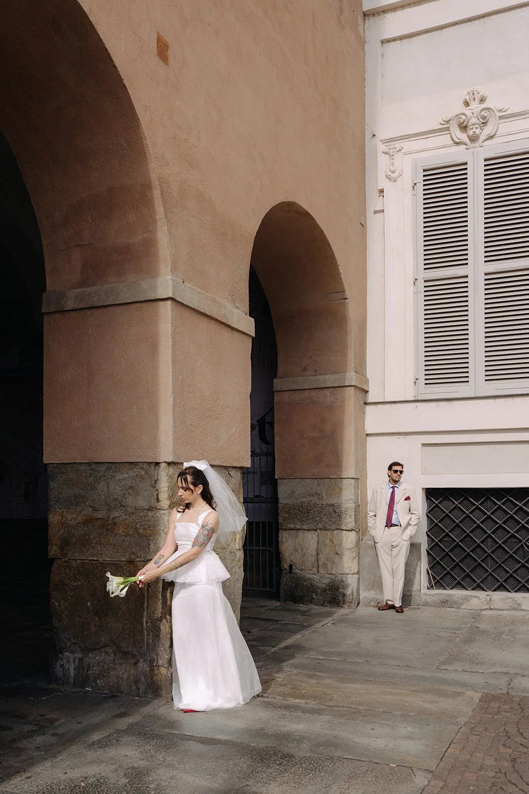 A woman in a white wedding dress holding a bouquet of flowers, standing near a stone archway, with a man in a light-colored suit and sunglasses leaning against the wall nearby.