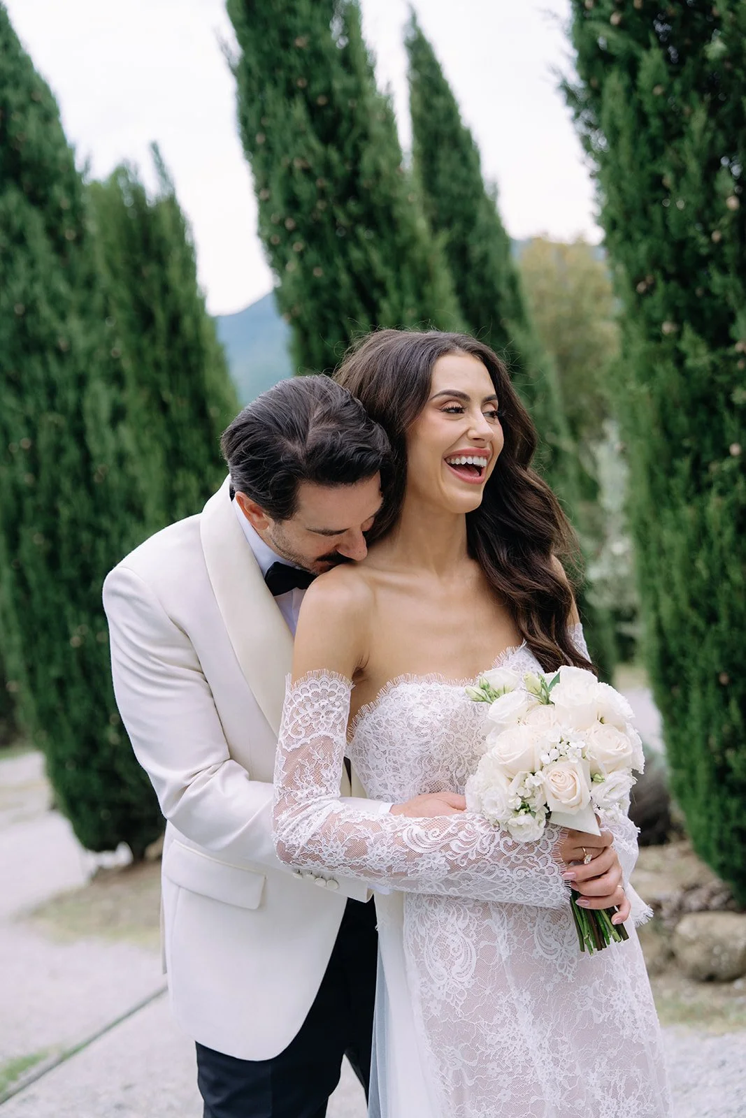 A happy bride in a lace wedding dress holding a bouquet of white roses, with a groom in a white tuxedo and black bowtie embracing her from behind in a garden with tall, green trees.