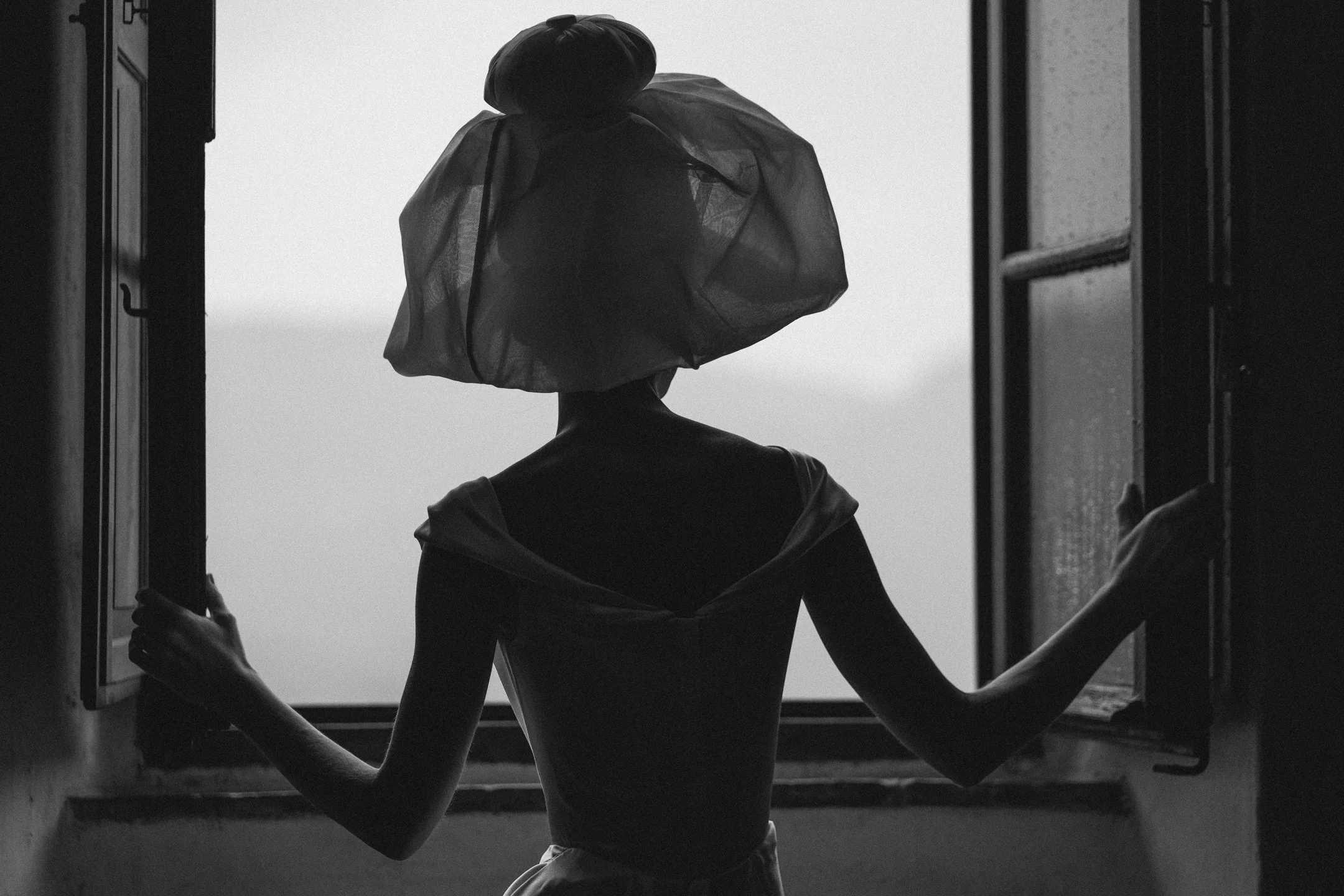 A person viewed from behind standing in front of an open window, wearing a large, transparent hat and elegant clothing, with arms extended to hold open the window shutters, in black and white.