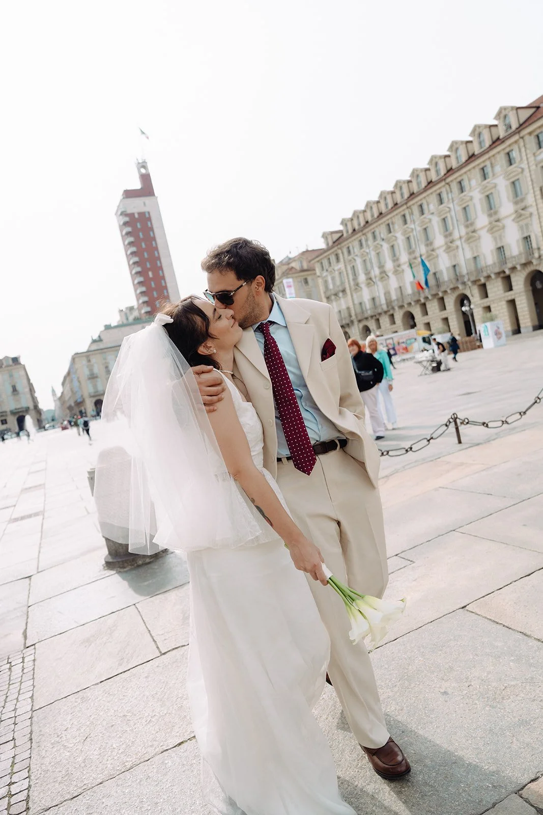 A bride and groom kissing outdoors, with the bride holding a bouquet of white calla lilies, in an urban setting with tall buildings and a clock tower in the background.