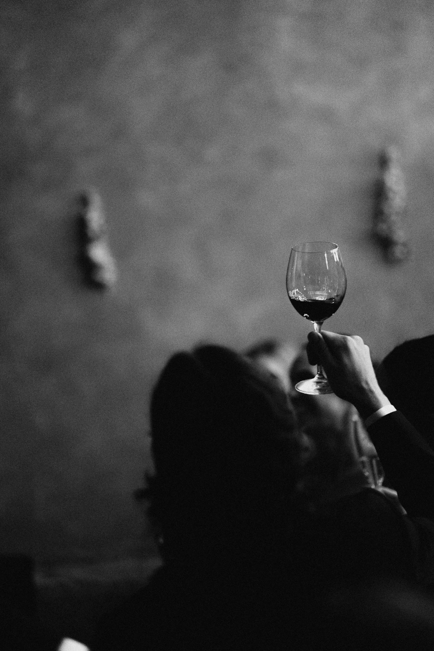 Person holding a glass of red wine in a dimly lit room, with blurred artwork on the wall in the background.