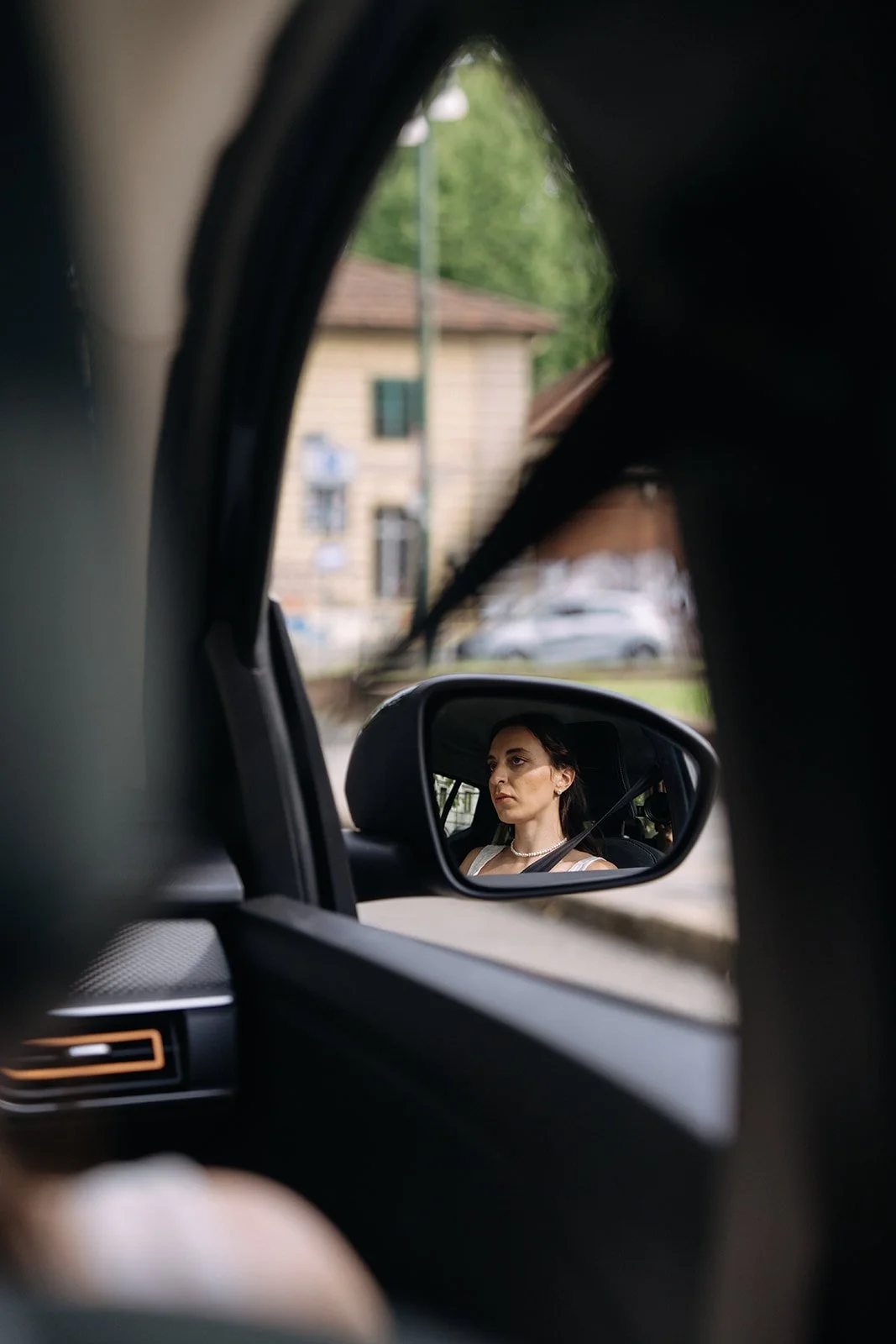 A woman wearing a white top and pearl necklace is seen through the side mirror of a car, with a house and parked cars in the background.