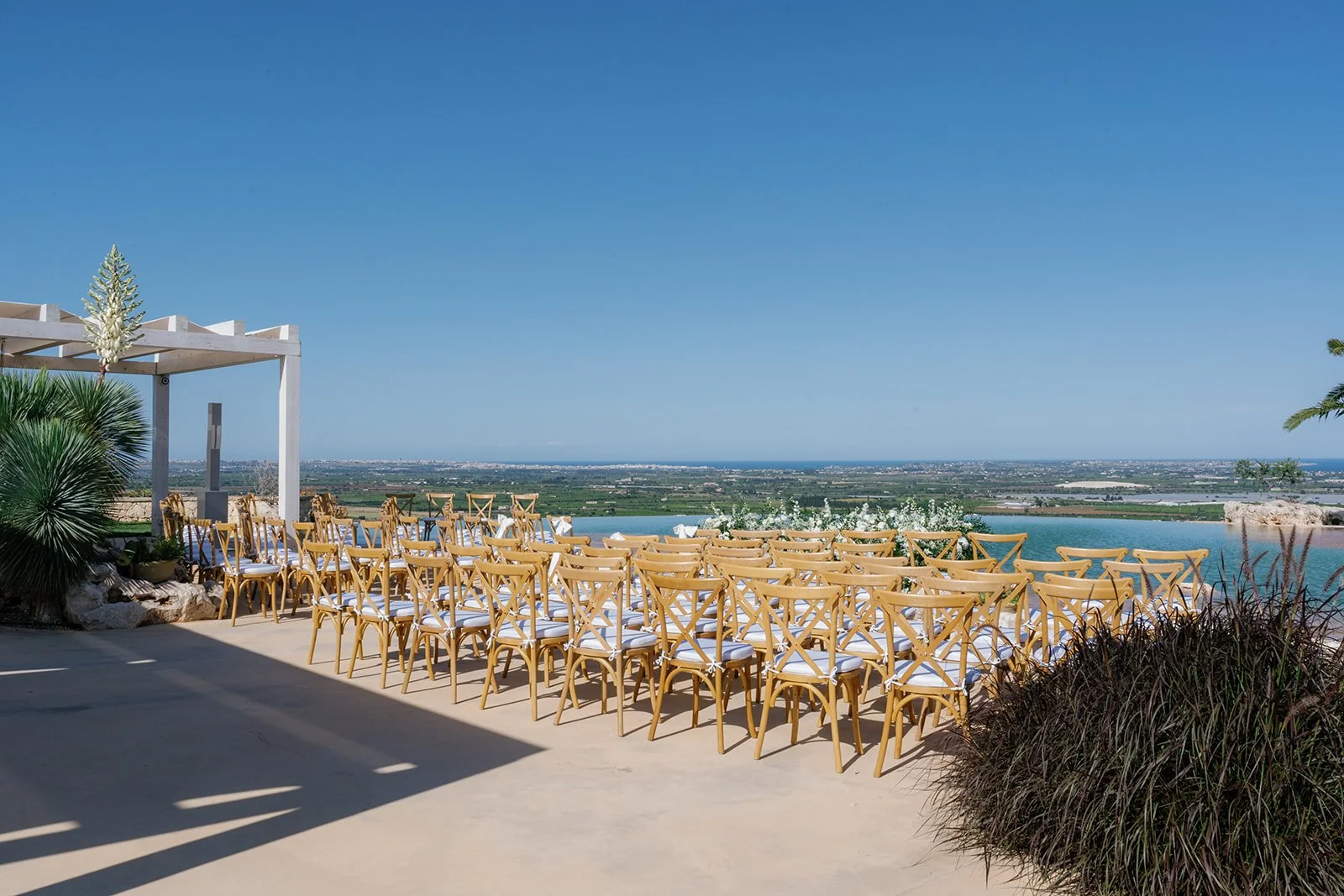 Outdoor wedding ceremony setup with rows of light wood chairs with white cushions facing an ocean view, under a white pergola with a beach landscape and clear blue sky in the background.