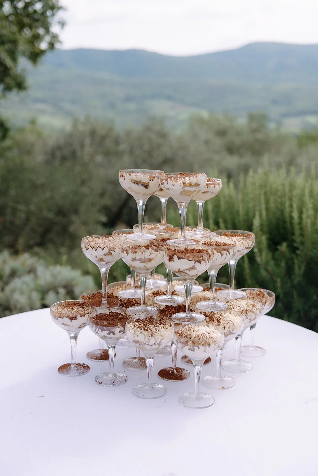 A pyramid of dessert glasses filled with layered tiramisu on a white table outdoors, with a blurred green landscape and mountains in the background.