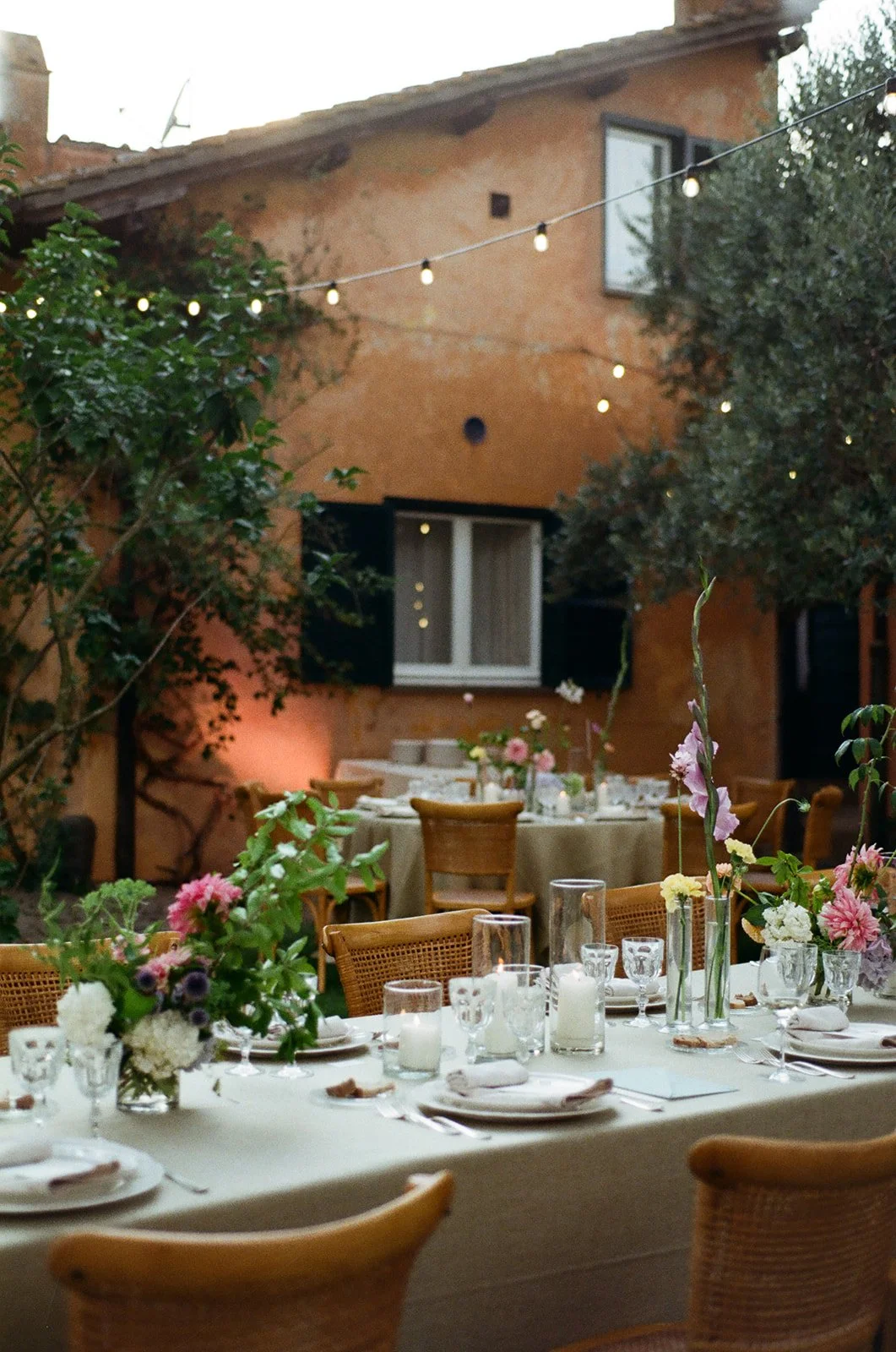 Outdoor dinner setup with decorated tables, floral centerpieces, string lights, and a rustic building in the background.