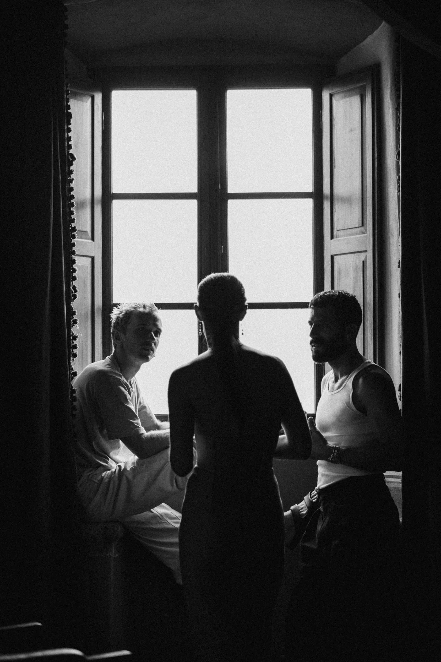 Silhouettes of three people — two men and one woman — sitting and standing by a large window, in black and white.