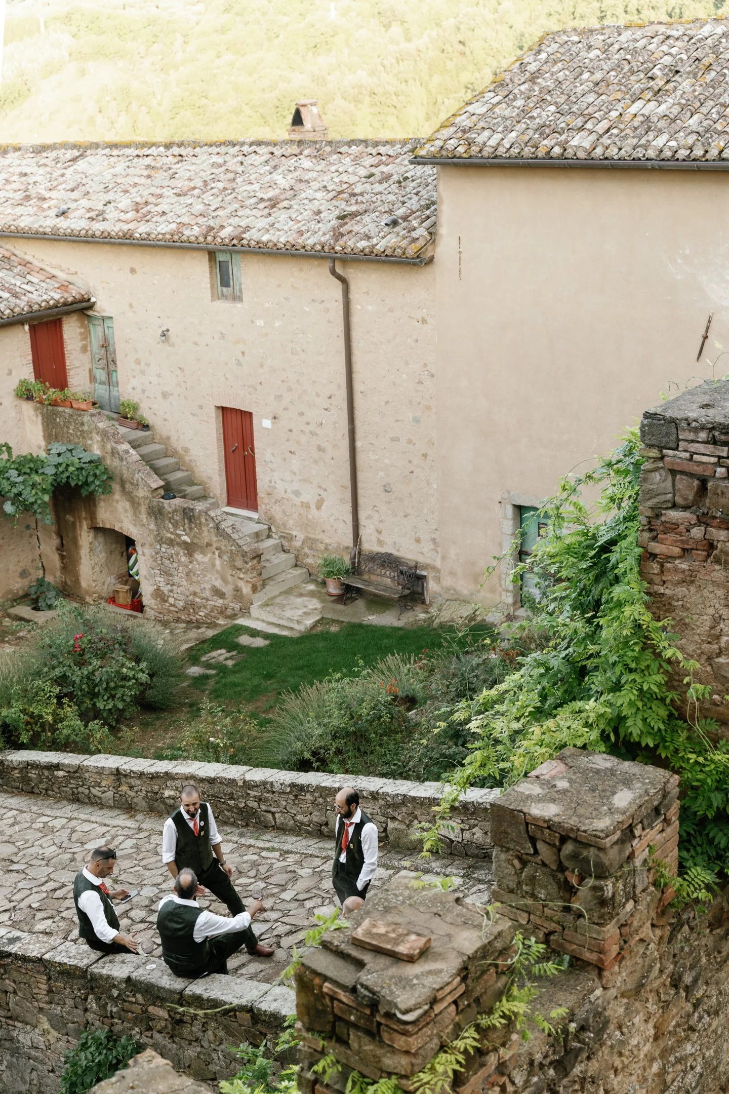 Four men dressed in traditional suits with vests and ties sitting and standing on a stone patio in a garden, having a conversation.