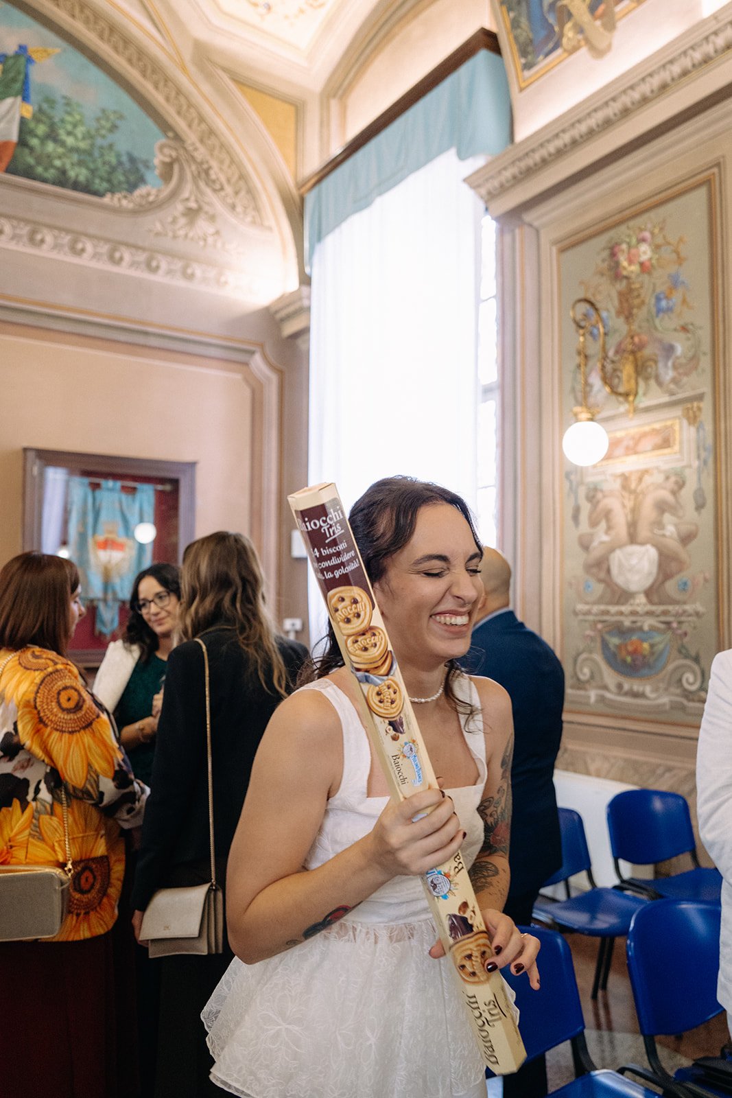 A woman in a white dress smiling and holding a long box of Italian cookies, surrounded by other people in a decorated room with ornate walls and high ceilings.