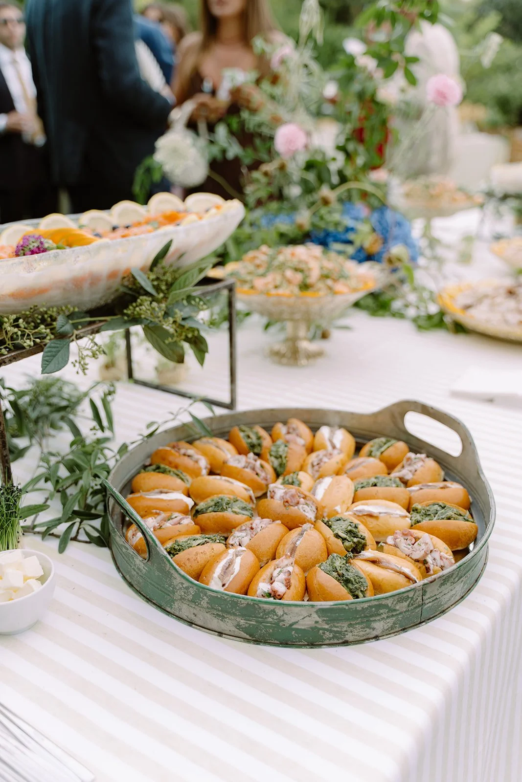 Tray of mini sandwiches filled with green and pink toppings on a striped tablecloth, decorated with greenery and flowers.