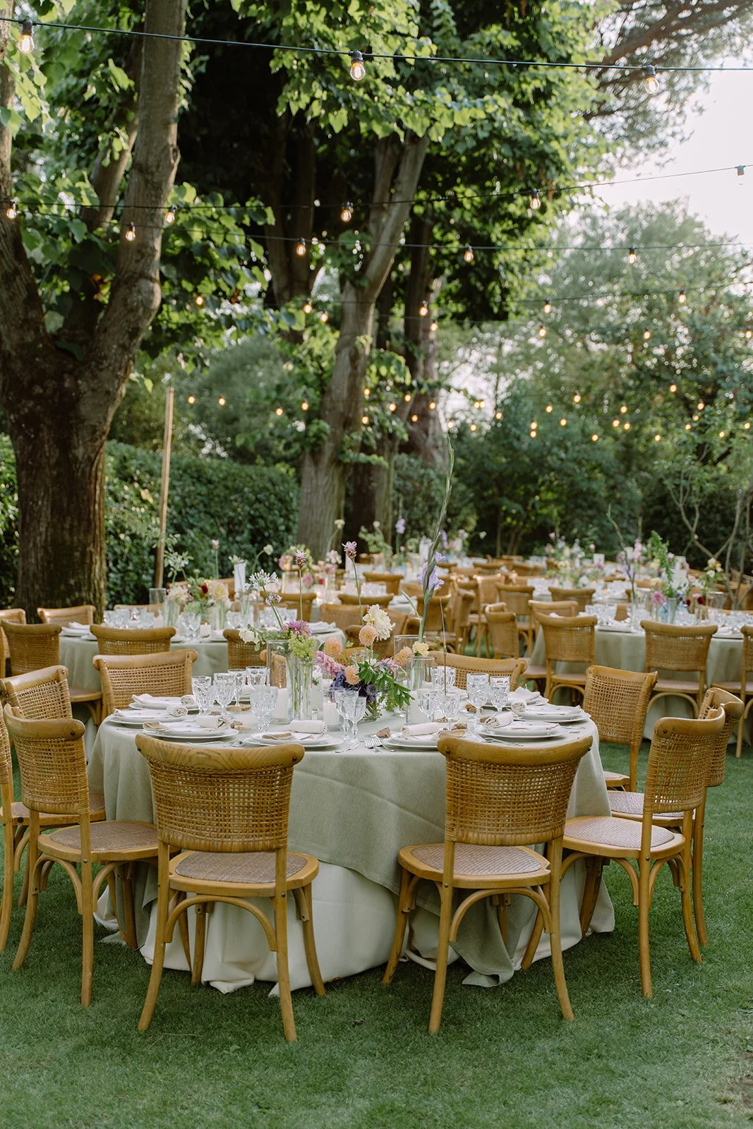 Outdoor wedding reception area with round tables, floral centerpieces, wicker chairs, and string lights hanging from trees. Green grass and foliage surround the setting.