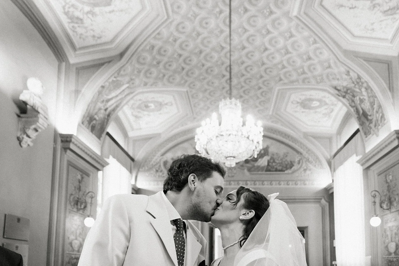 A black and white photo of a newlywed couple sharing a kiss in an ornately decorated hall with a chandelier overhead.