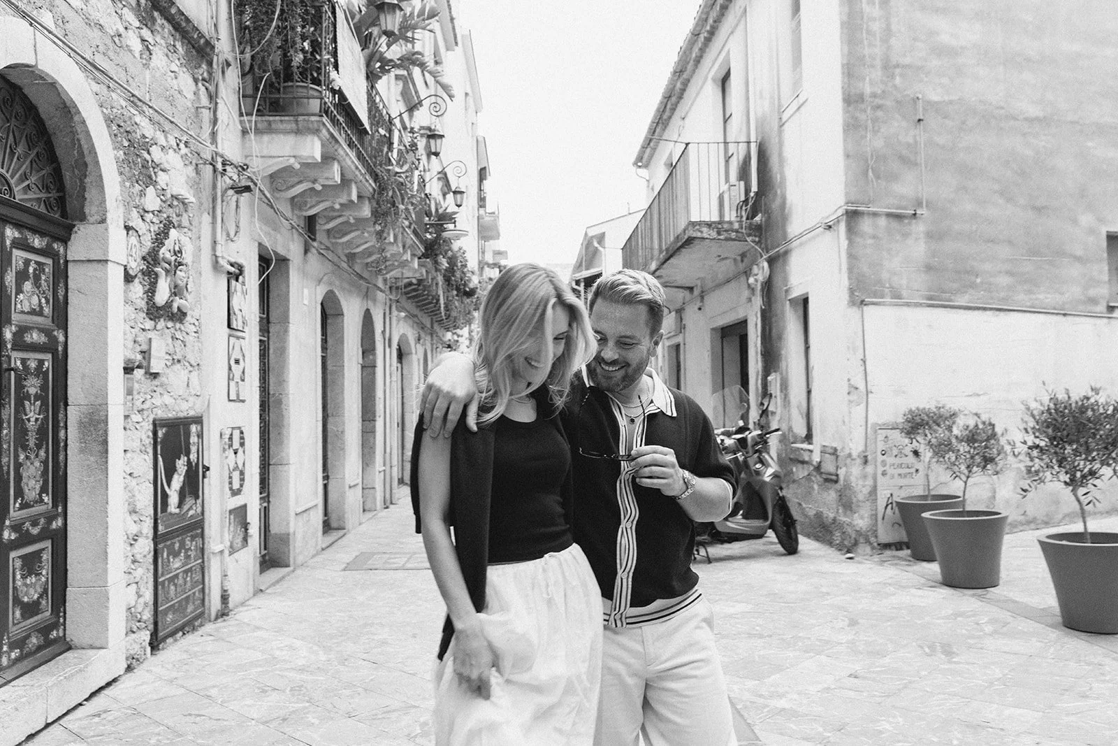 A man and woman walking together on a narrow outdoor city street, smiling and enjoying each other's company, with old buildings and potted plants in the background.