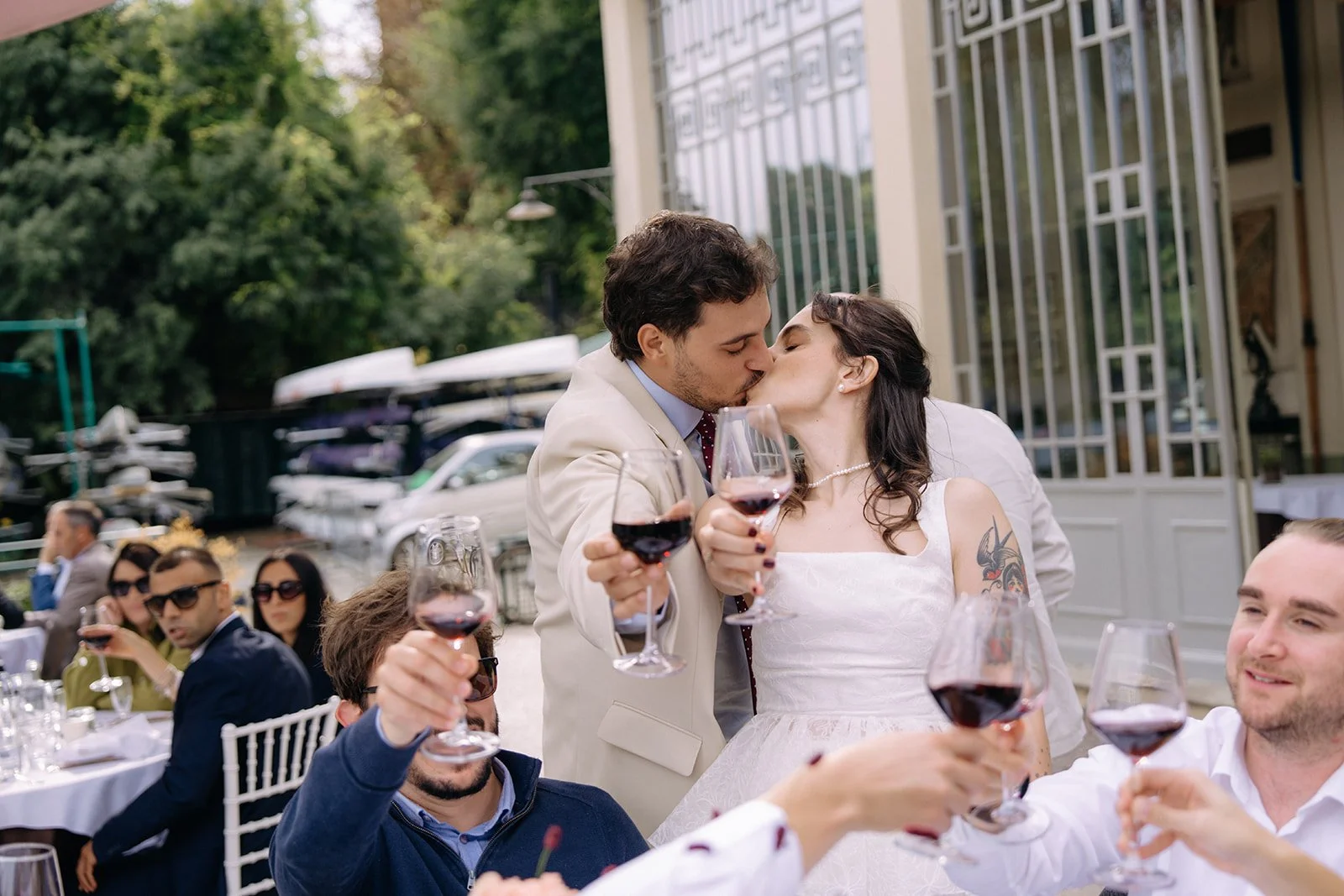 A couple in wedding attire kissing at a celebratory outdoor event, with guests raising glasses of red wine