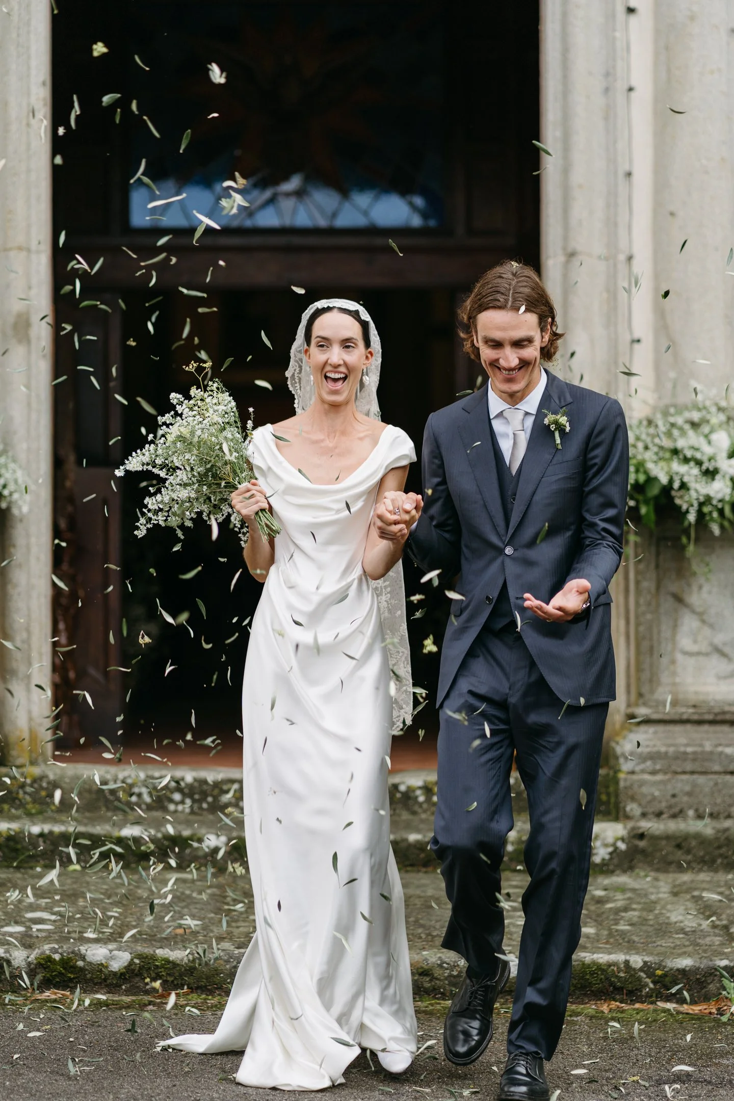 A newly married couple walks out of a building, celebrating with falling confetti. The bride is dressed in a white wedding gown holding a bouquet, and the groom is in a navy suit. They are smiling and holding hands.
