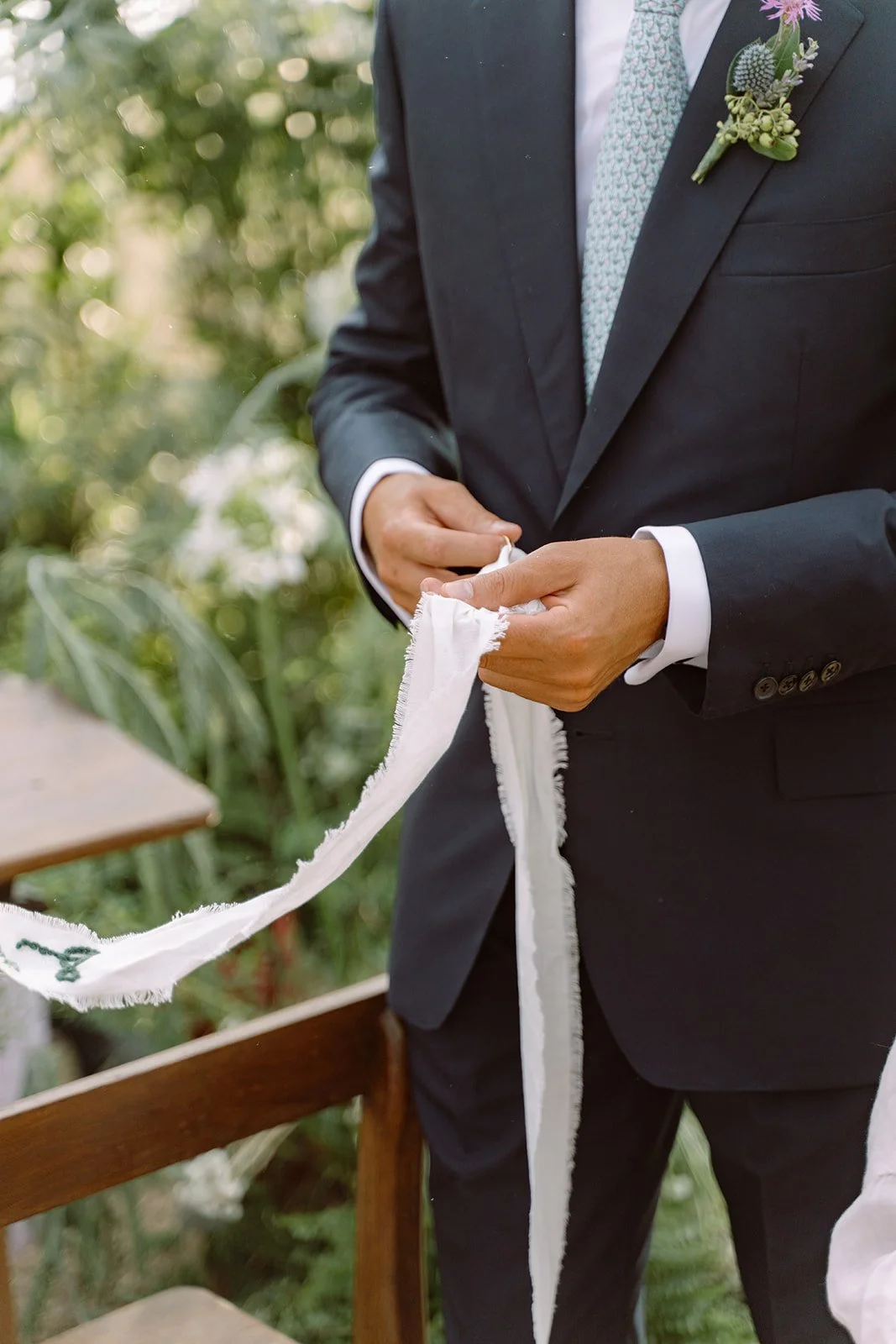 A man dressed in a dark suit and tie is holding a white cloth ribbon during a wedding ceremony outdoors.