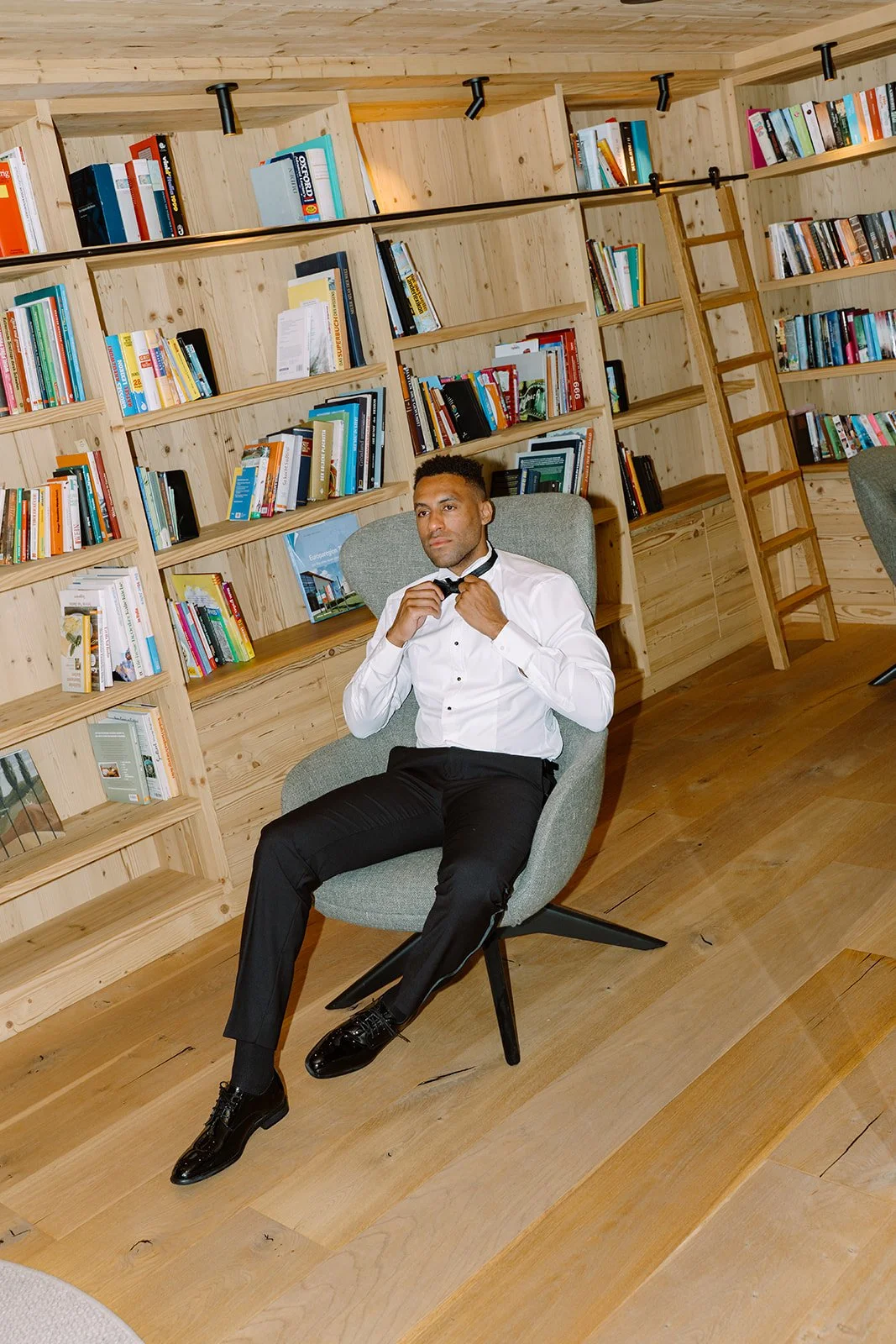 A man in a white dress shirt and black pants sitting in a modern library or study room, adjusting his tie while seated in a grey armchair. The background features wooden bookshelves filled with colorful books and a small ladder.