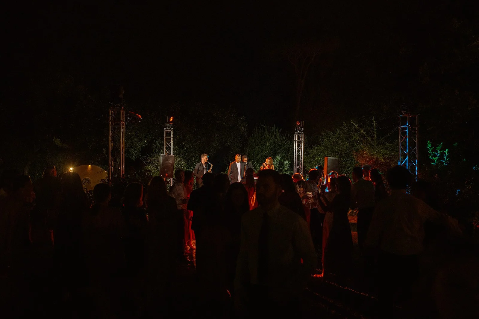 People dancing and socializing outdoors at night, with a band performing on a stage under colorful lights and trees in the background.
