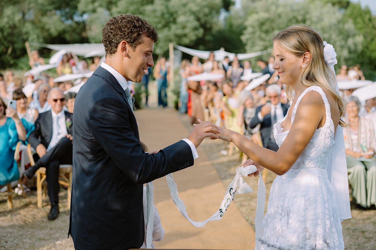A couple exchanges wedding rings during an outdoor ceremony with guests seated and standing under parasols in the background.