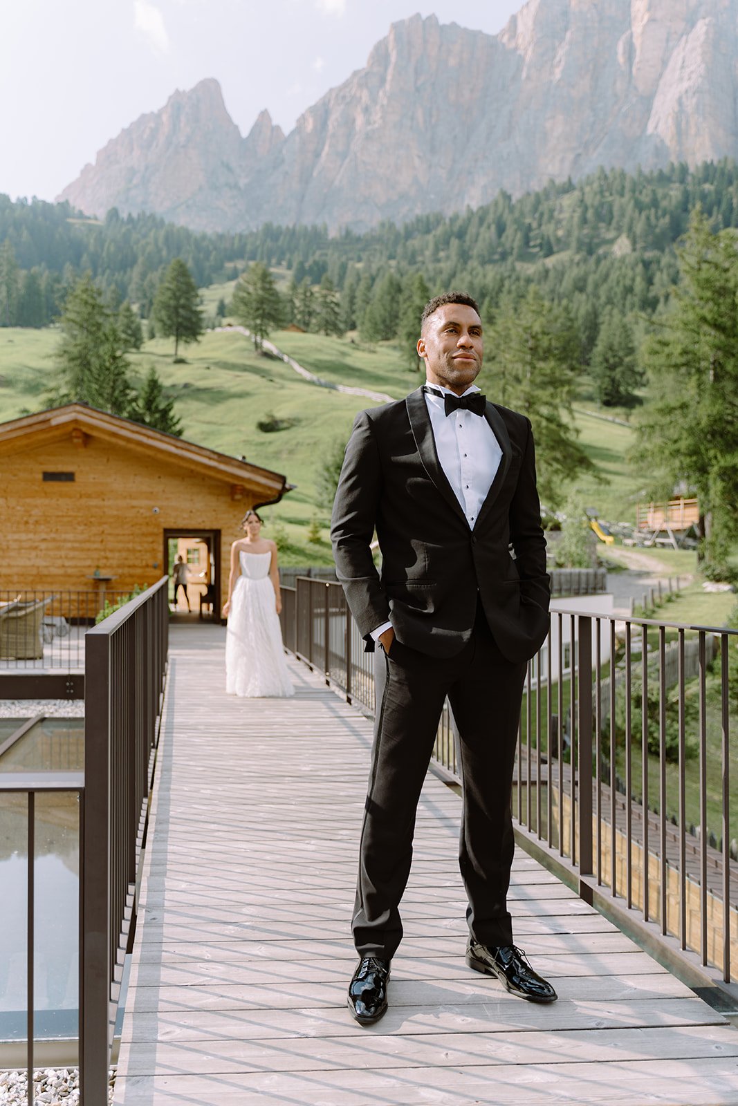 A groom in a black tuxedo with a bow tie stands on a wooden bridge with hands in pockets, looking into the distance. A bride in a white wedding dress stands behind him, looking on. The scene is outdoors with lush green trees, mountains, and a wooden 