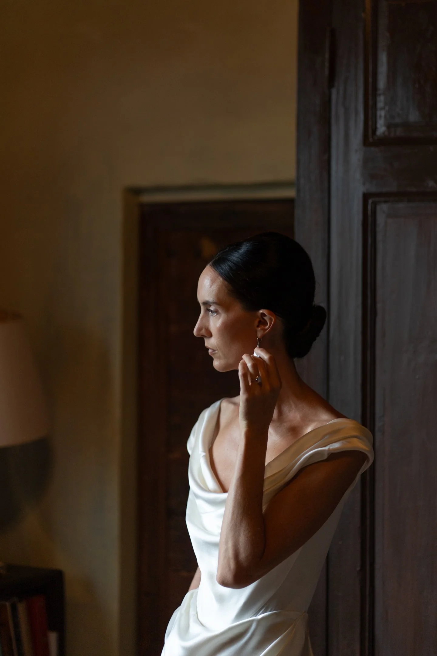 A woman with dark hair styled in a bun, wearing a white dress, stands in profile indoors in front of a dark wooden door, touching her earring.