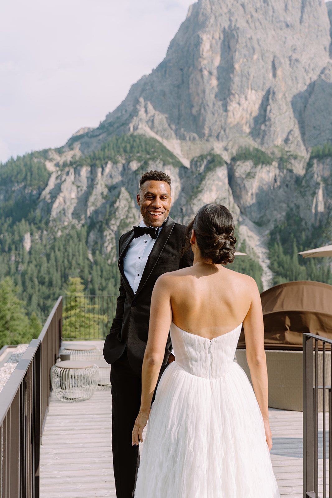 A newlywed couple standing on a wooden deck outdoors with a mountain landscape in the background. The groom is wearing a tuxedo and smiling at the bride, who is dressed in a strapless white wedding gown with her back to the camera.