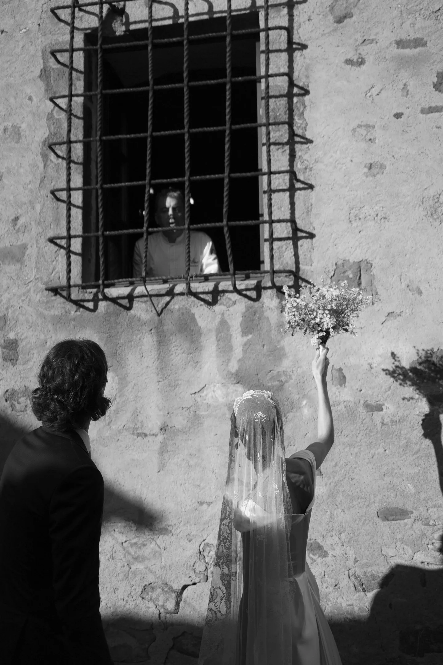 A woman in a wedding dress and veil holds a bouquet, standing outside a stone building with a barred window. She faces a man in a suit, and a man inside the building looks out from the barred window.