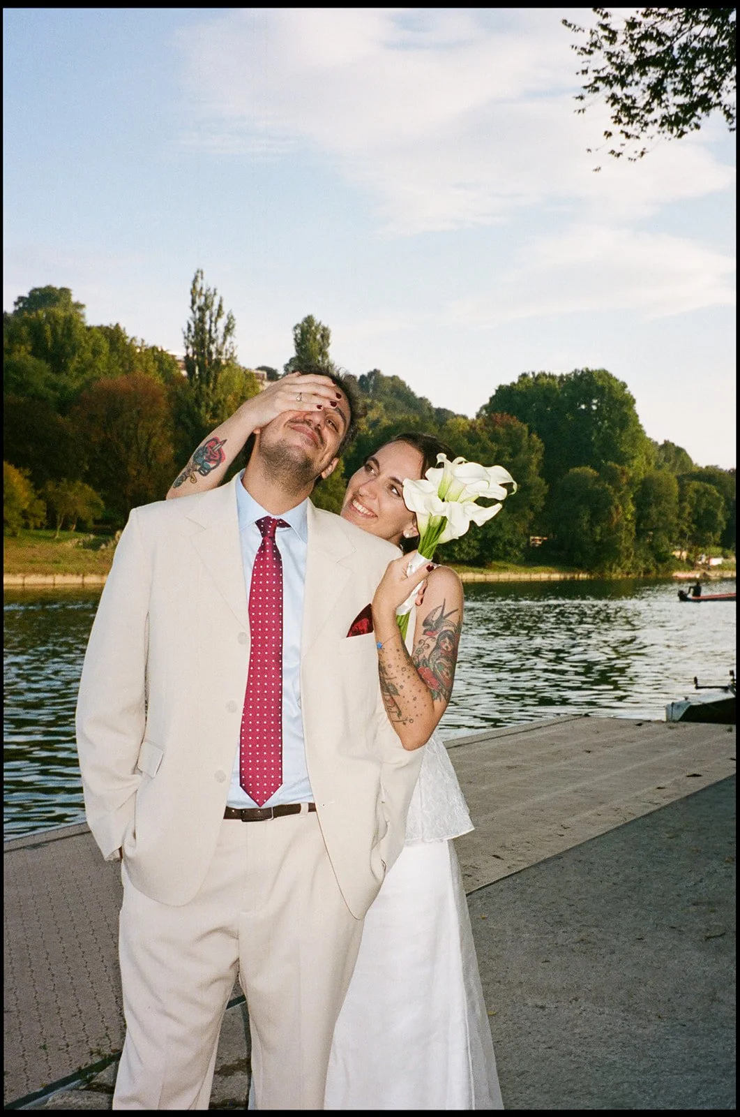 A happy couple standing by a river with trees in the background. The woman is holding a bouquet of white calla lilies and smiling at the man. The man is dressed in a cream-colored suit with a red polka-dot tie, and the woman has tattoos on her arms a