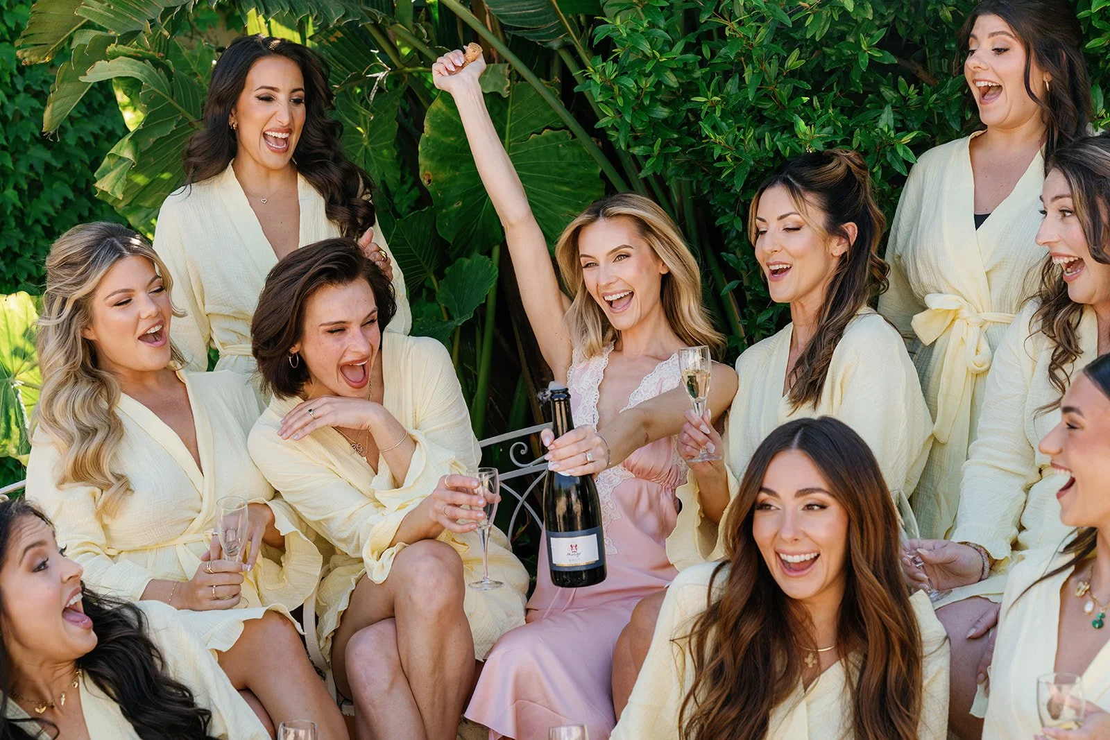 A group of women celebrating outdoors, wearing matching yellow robes, with one woman in a pink dress holding a champagne bottle and glass, surrounded by lush green foliage.