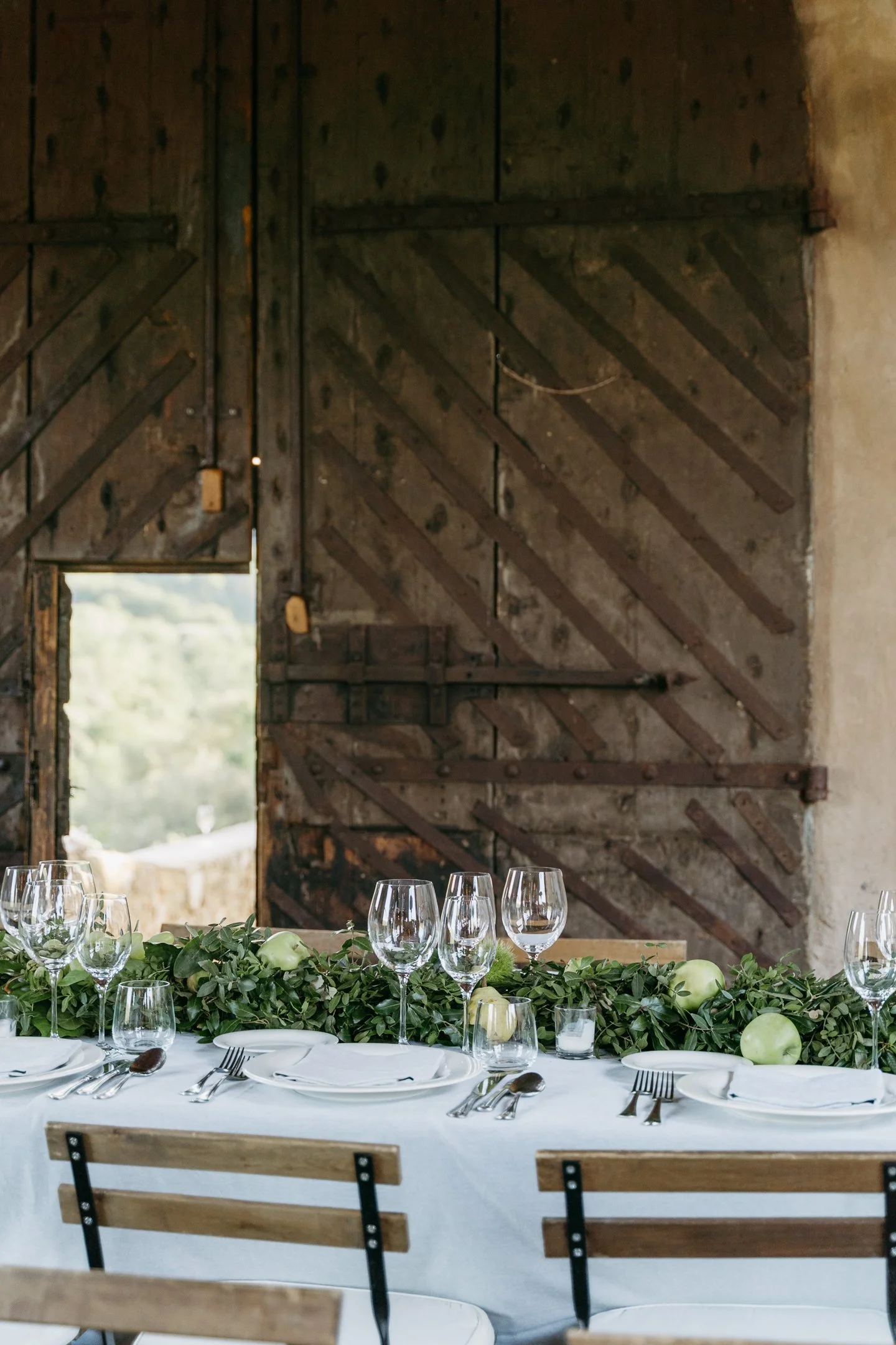 A rustic dining table set for a meal with a green foliage centerpiece and white apples, in front of a weathered wooden barn door with a small window opening.