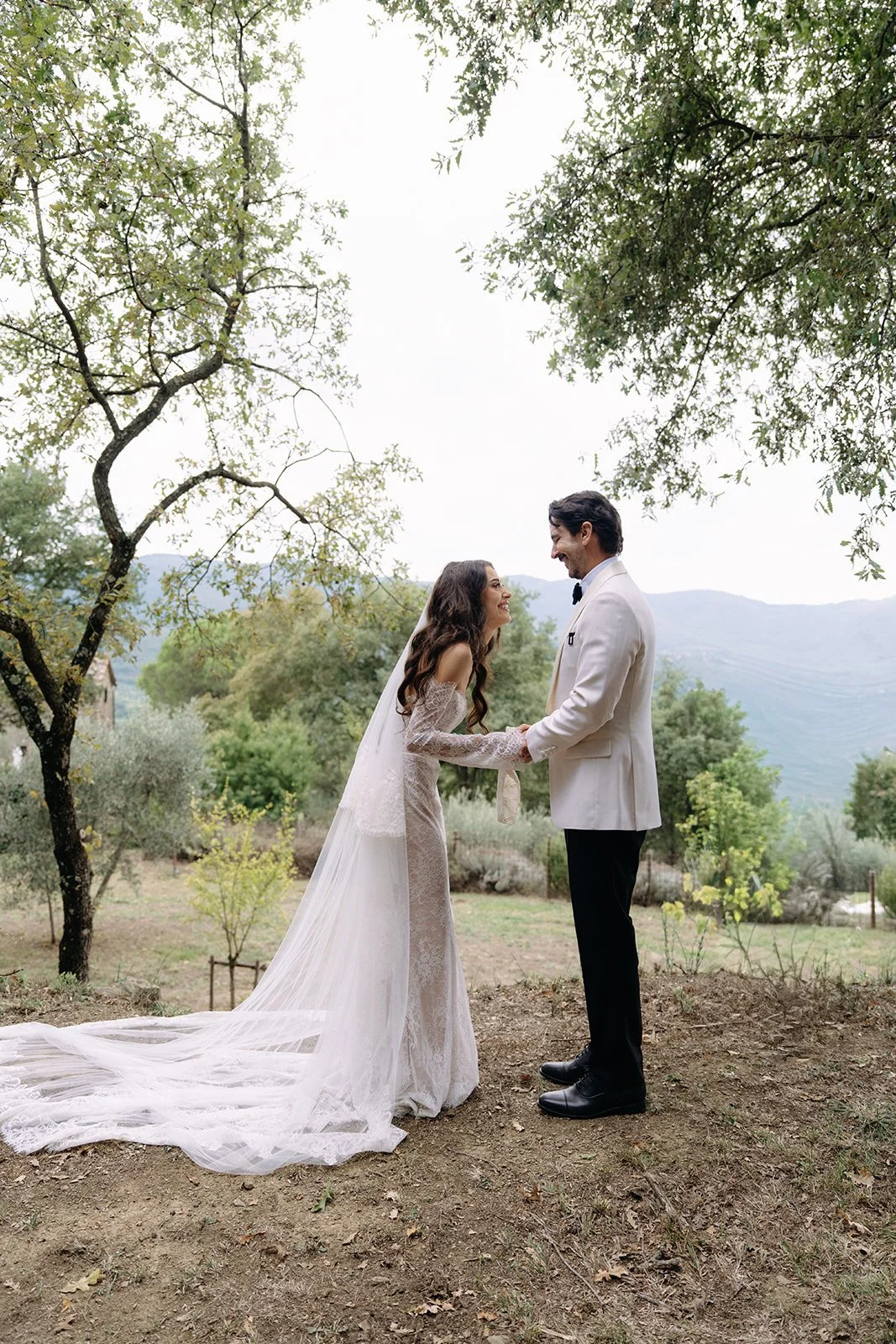 A bride and groom holding hands and smiling at each other outdoors with trees and mountains in the background.